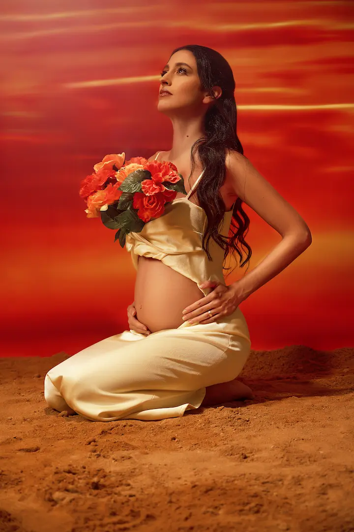Pregnant woman kneeling gently on the sand, cradling vibrant red flowers over her belly, looking upwards with a peaceful glow, embraced by the warmth of a red sunset sky, symbolizing life, love, and transformation