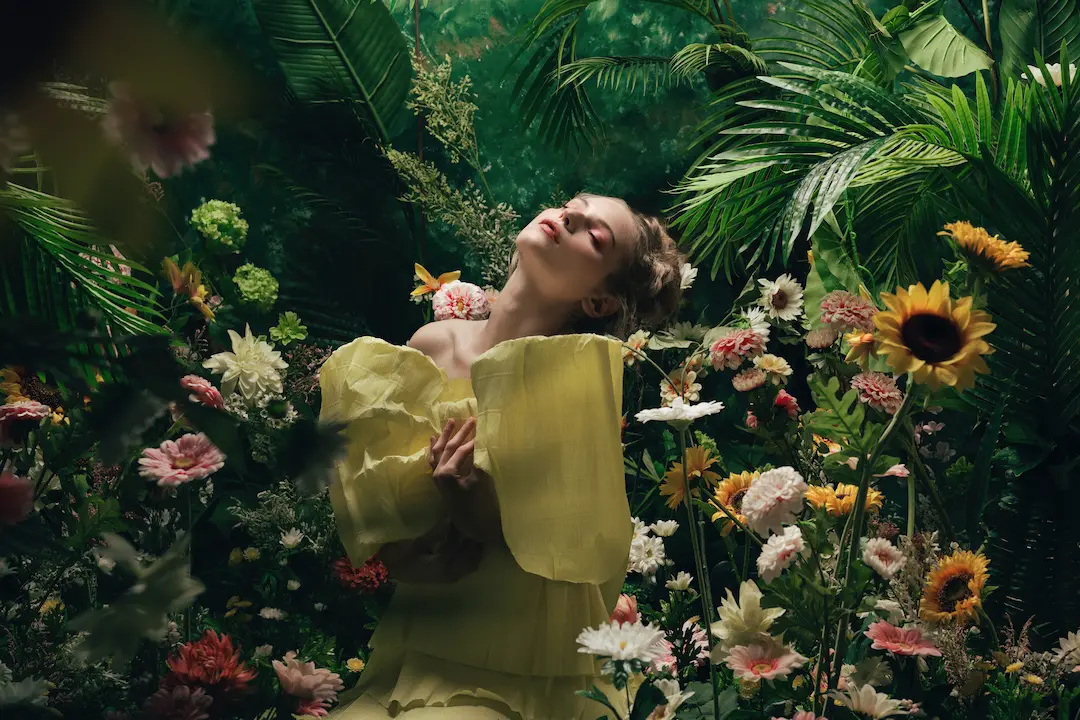 Woman in a yellow dress surrounded by vibrant flowers and lush green foliage, posing with her eyes closed in a serene and natural environment